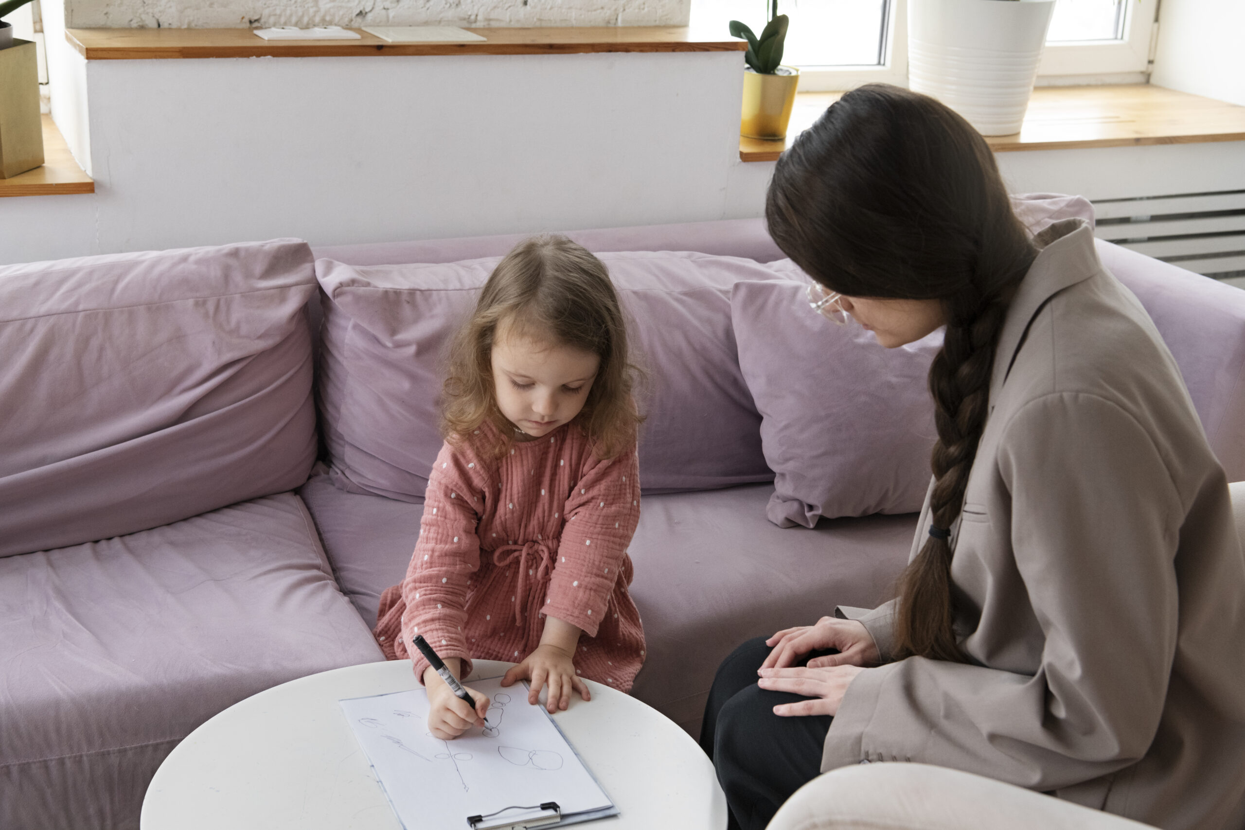 A young girl draws on paper at a round white table while an adult with long brown hair watches her in a cozy room with a light purple sofa and potted plants.