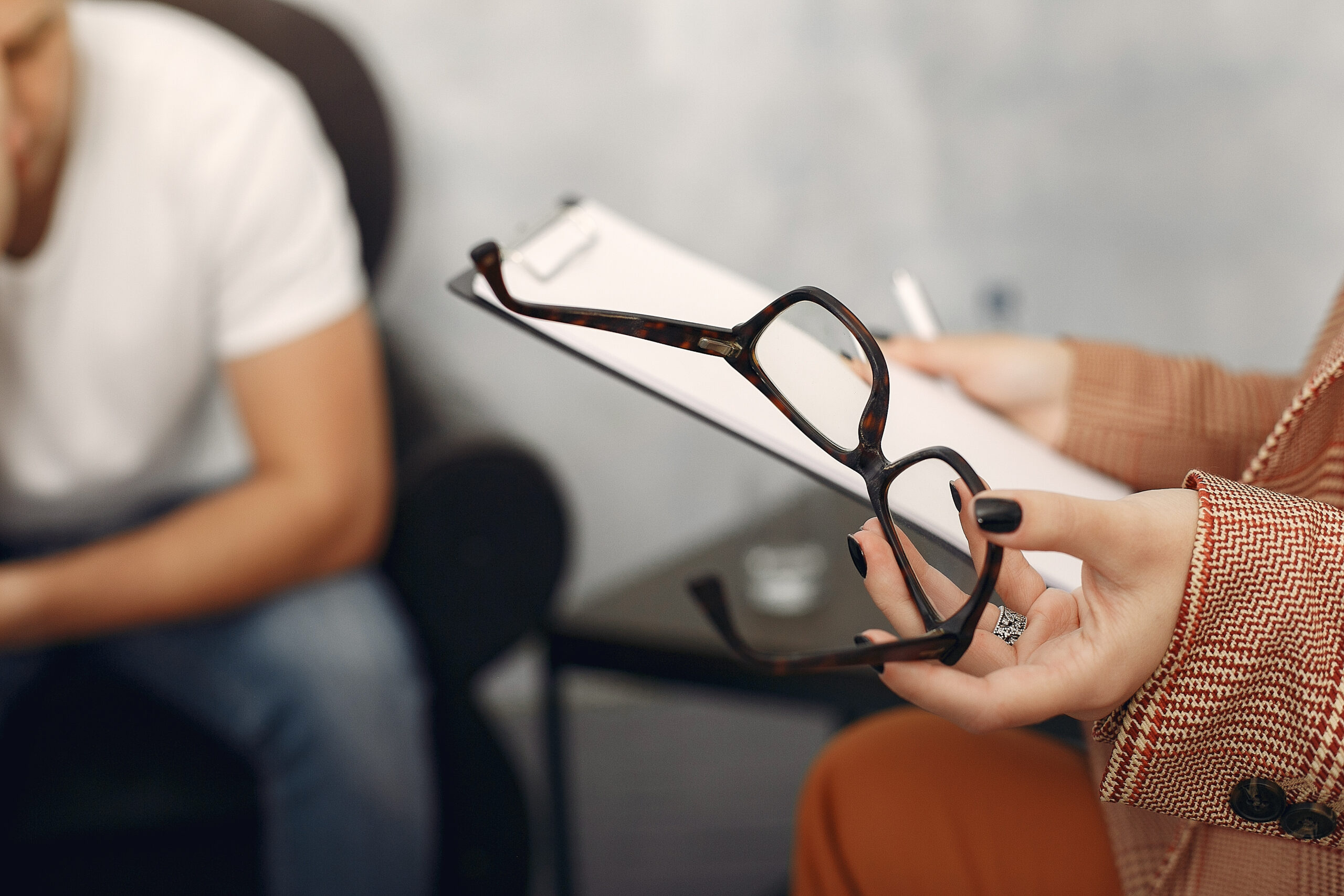 A person holding eyeglasses and a clipboard with a pen, possibly a therapist or counselor, sits across from another person in a white shirt, who appears to be in conversation or deep thought.