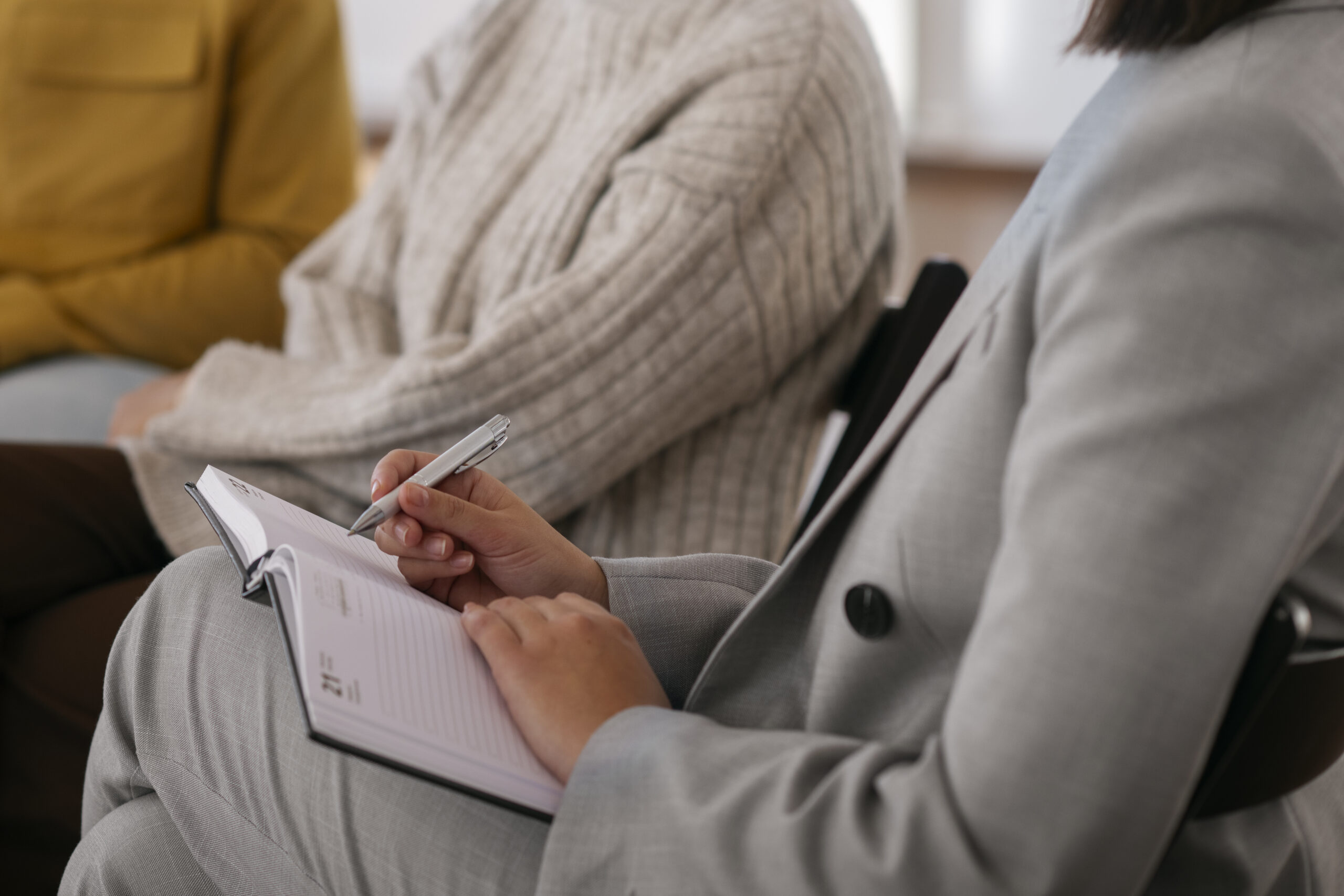 Three people sit side by side; one in a gray suit writes in a notebook with a pen, while the others wear casual sweaters. The image focuses on their torsos and hands, not their faces.