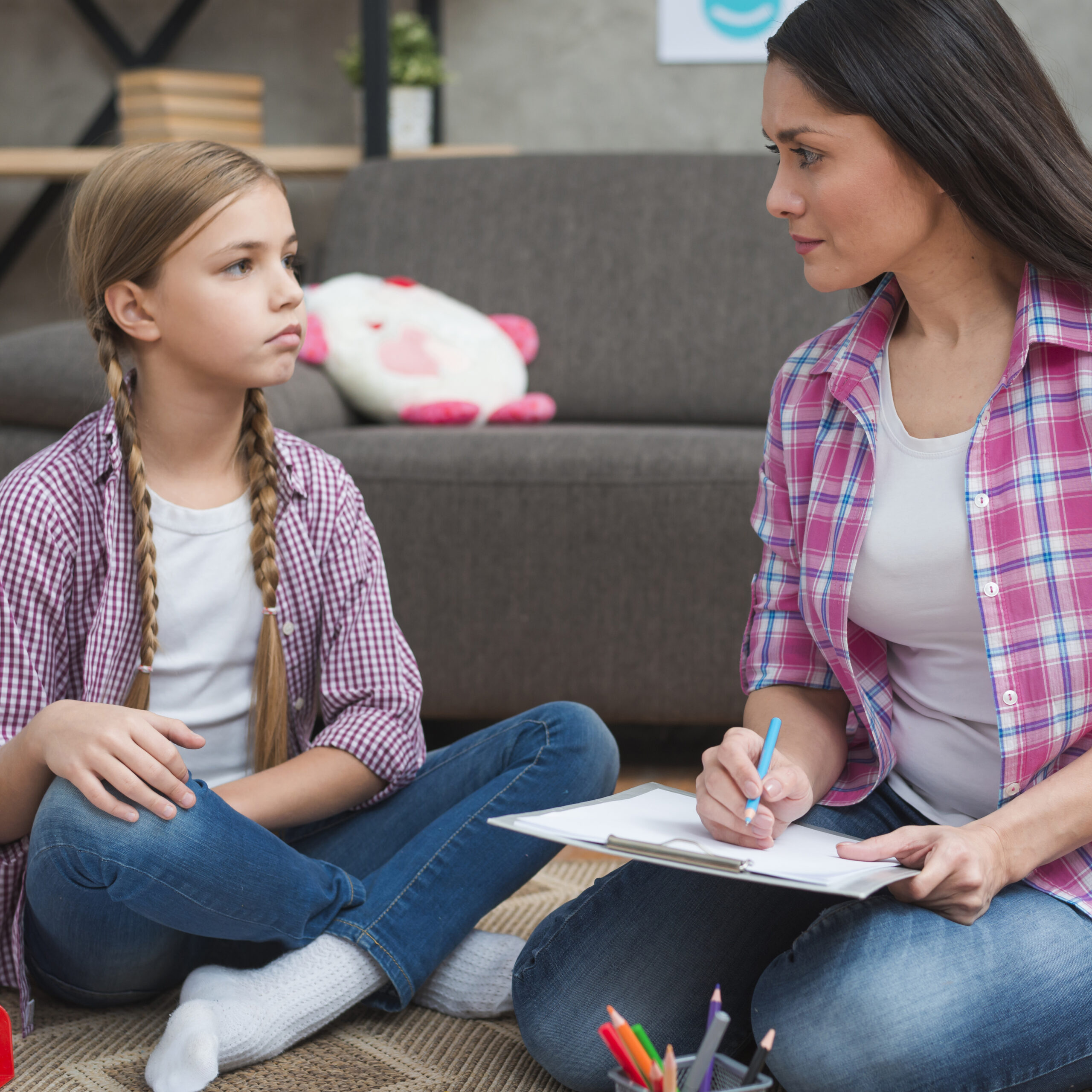 A woman with a clipboard sits cross-legged on the floor facing a young girl with braided hair. Both wear plaid shirts and jeans, suggesting a conversation or counseling session in a cozy room with toys and a sofa in the background.