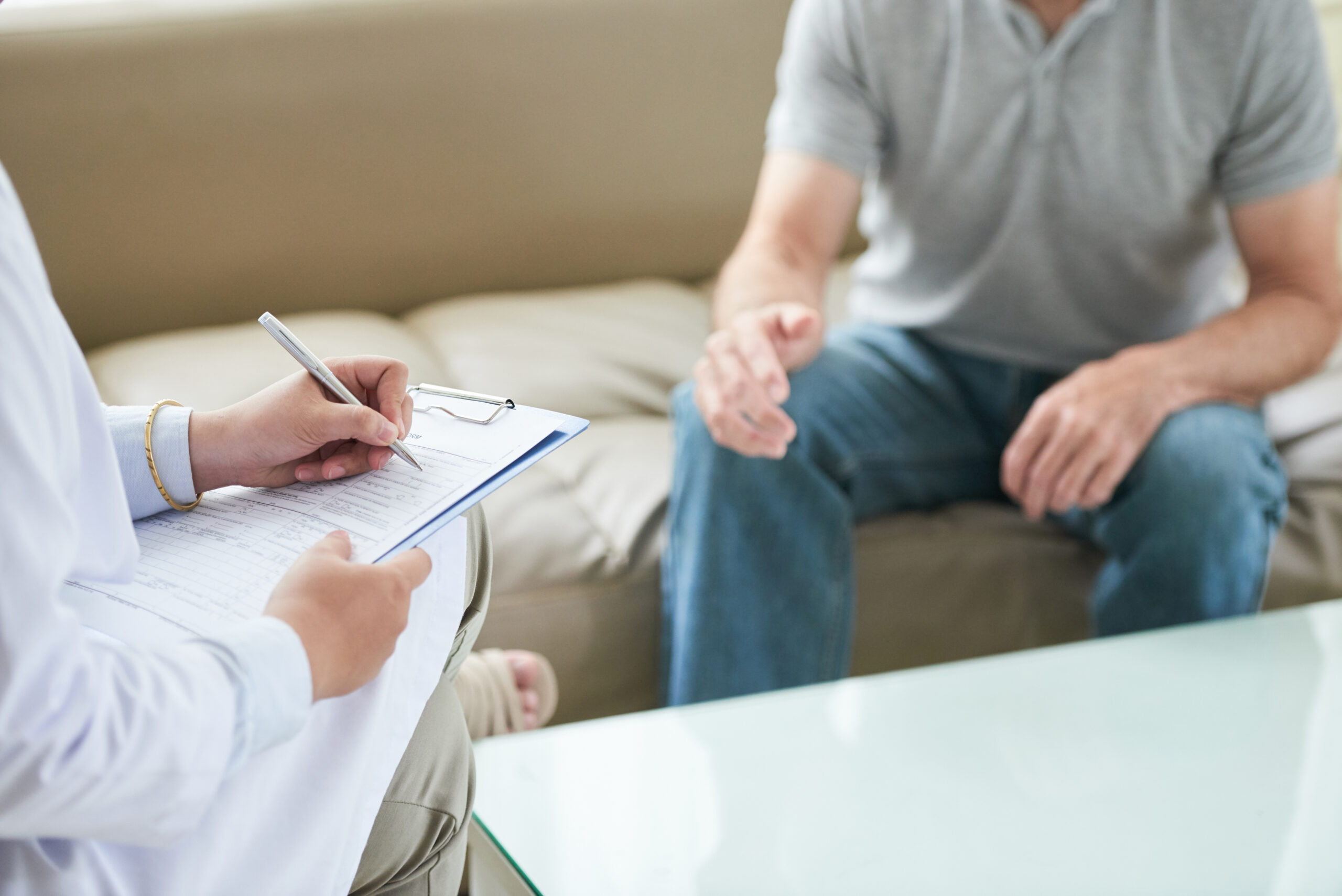 A person in a white coat writes on a clipboard, while another person in casual clothes sits on a couch, gesturing with one hand during a conversation.